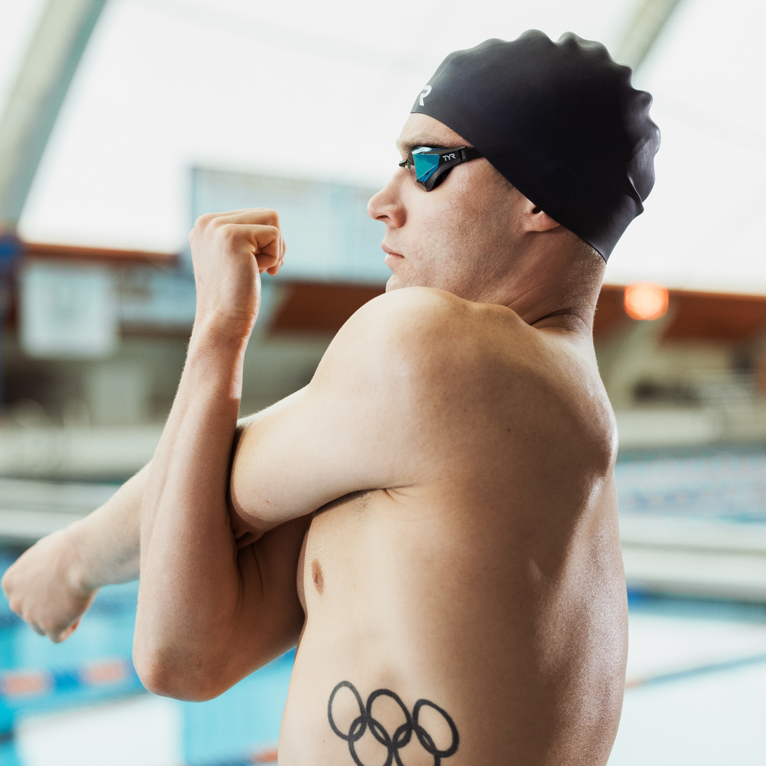 Man wearing a swim cap and goggles with an Olympic ring tattoo, standing by a pool.