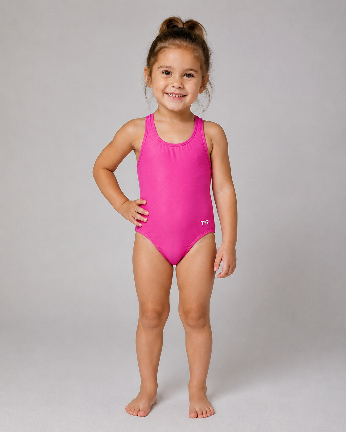 A young girl with her hair in a high ponytail stands barefoot against a light gray studio background, wearing a TYR bright pink one-piece swimsuit. The swimsuit has a scoop neckline and a small white TYR logo on the lower right hip.